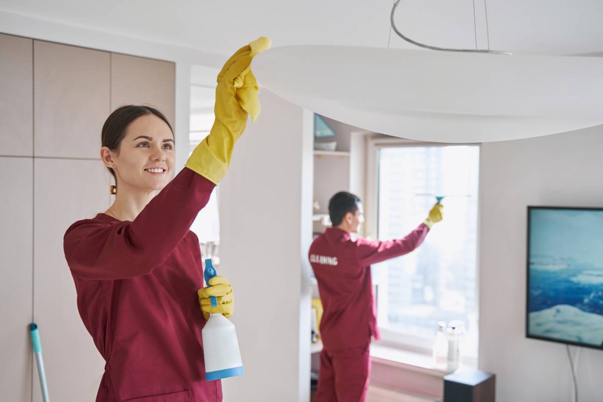 Smiling janitor wiping uplight bowl pendant with microfiber cloth while her coworker washing window