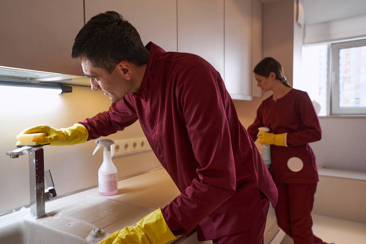 Janitor wiping chrome faucet with sponge and detergent while his colleague cleansing countertop