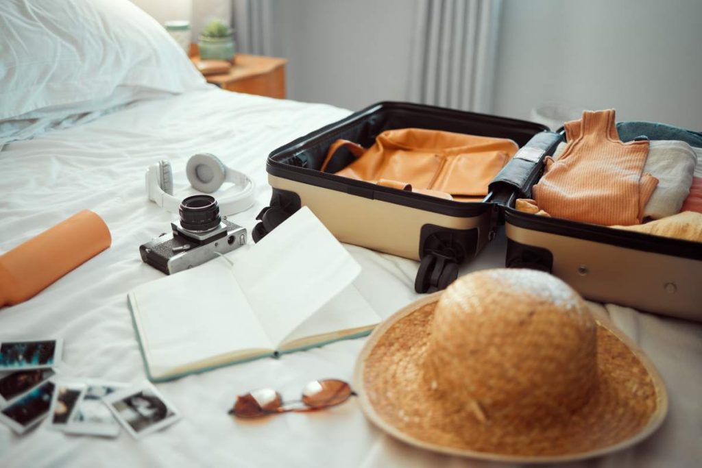 Travel, luggage and bedroom with a suitcase, hat and camera on a bed in a hotel during holiday or v.