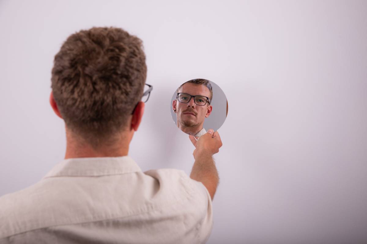 Serious caucasian man looks at himself in the mirror and reflects on life. Pensive person with eyeglasses. Guy isolated on white background, Studio photo.